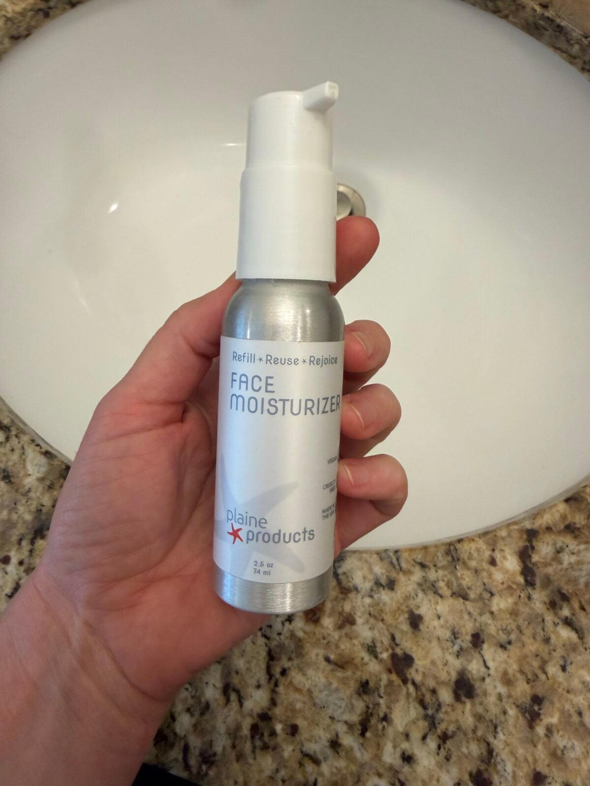 A hand holds a silver bottle of Plaine Products face moisturizer with a pump dispenser over a granite countertop and a white sink.