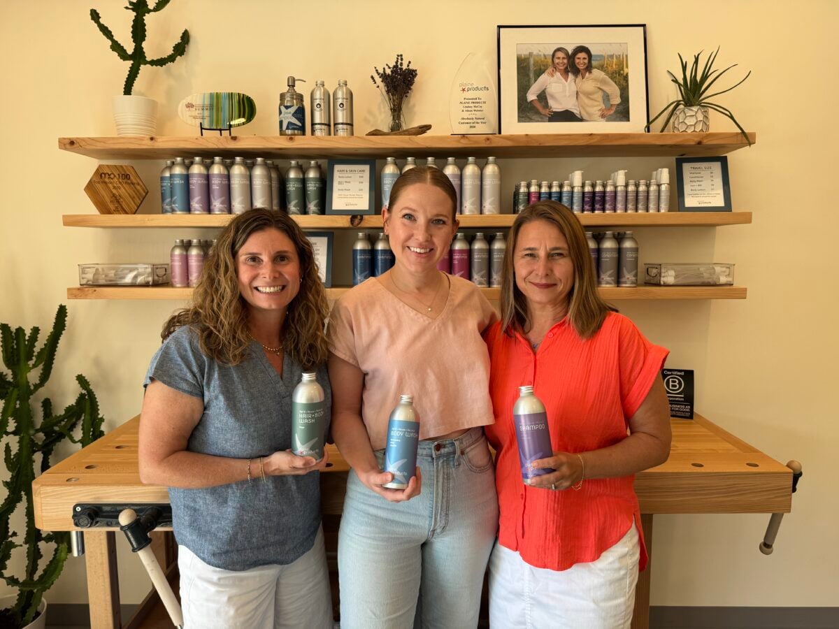 Three women stand in front of shelves with bottles, each holding a different colored bottle. The background features a framed photo, certificates, and plants, with a wooden table in front of the women.
