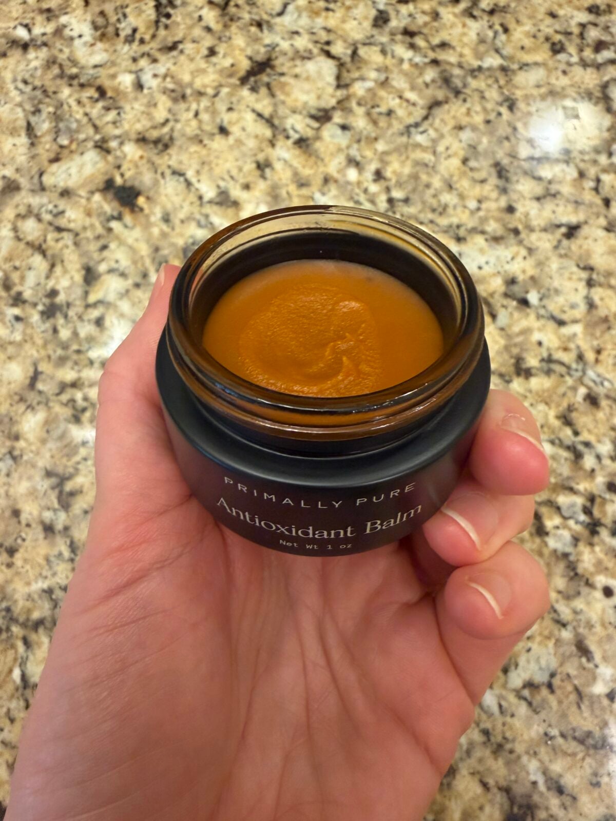 A hand holds an open jar labeled Primally Pure Antioxidant Balm against a speckled granite countertop. The balm inside is smooth and orange-brown in color.