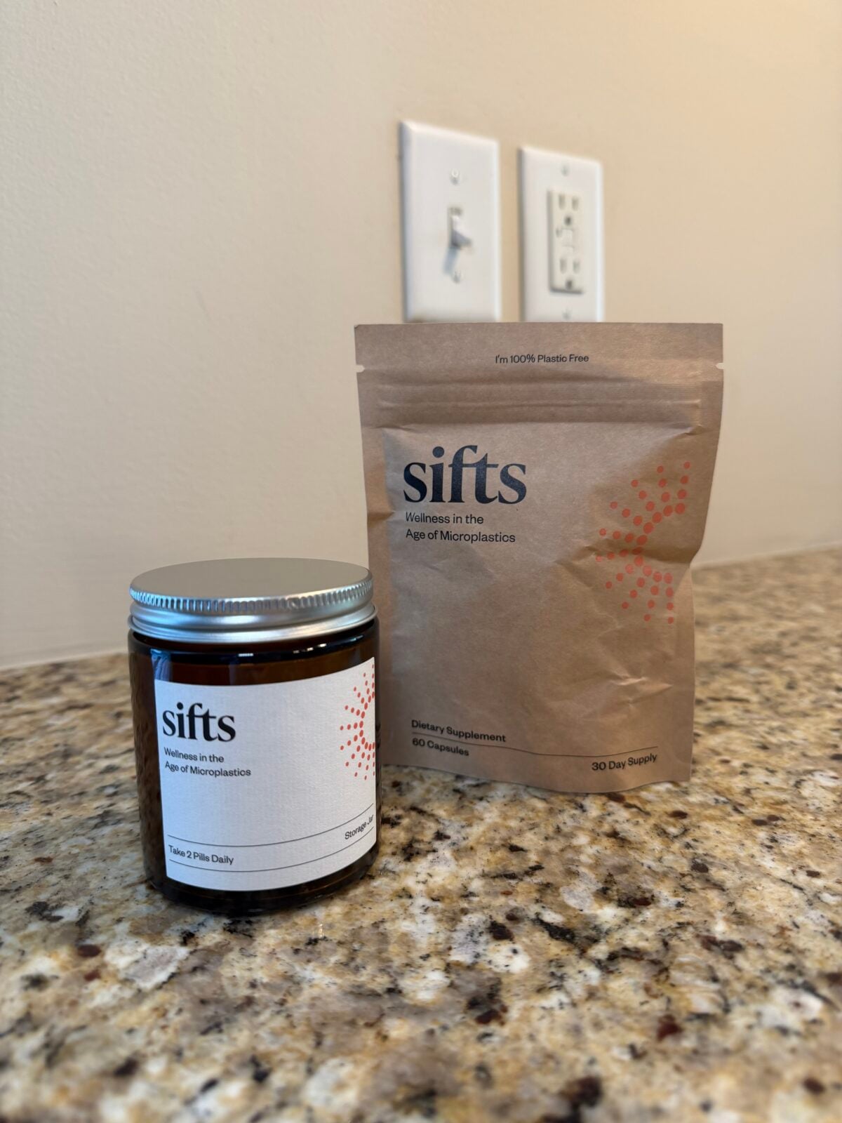 A brown glass jar and a brown paper pouch, both labeled sifts: Wellness in the Age of Microplastics, sit on a granite countertop. The jar has a metal lid; the pouch stands upright. Electrical outlets are visible on the wall behind.