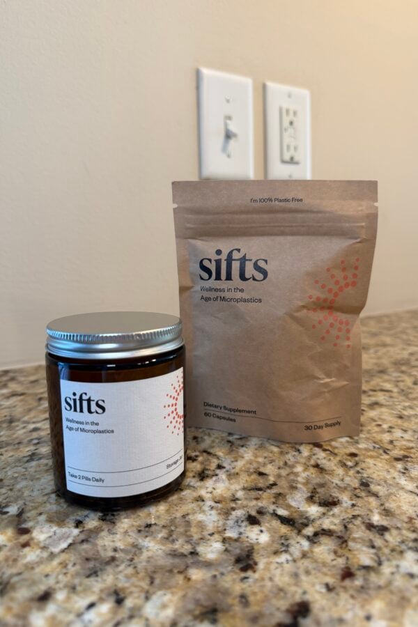 A brown glass jar and a brown paper pouch, both labeled sifts: Wellness in the Age of Microplastics, sit on a granite countertop. The jar has a metal lid; the pouch stands upright. Electrical outlets are visible on the wall behind.