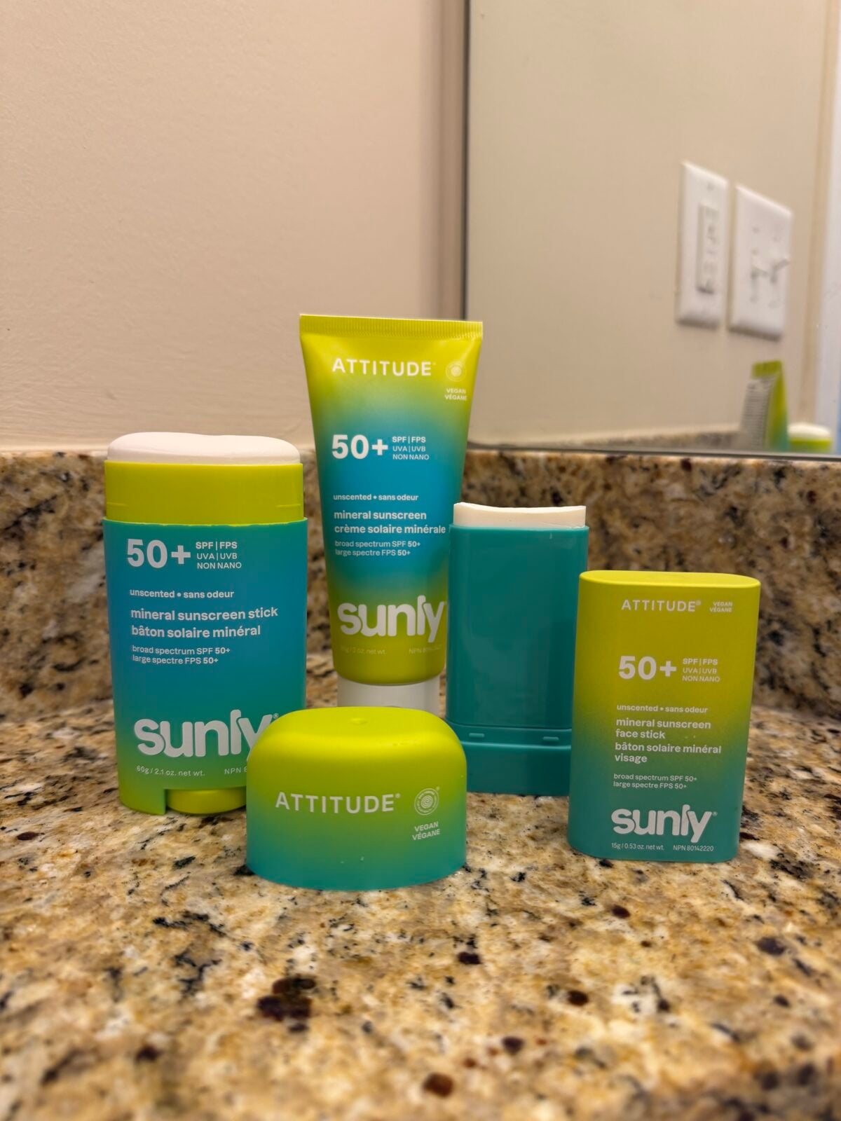 Five Attitude Sunly mineral sunscreen products are arranged on a bathroom counter with a granite surface; two tubes, two sticks, and one container are visible, all labeled SPF 50+ with blue and green packaging. A mirror reflects part of the scene.