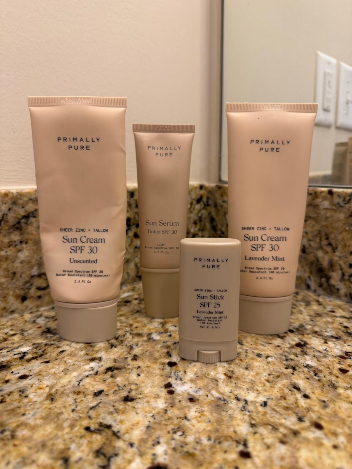Four Primally Pure sunscreen products, including two tubes of Sun Cream SPF 30, one Sun Serum Tinted SPF 30, and one Sun Stick SPF 25, are lined up on a granite bathroom counter in front of a mirror.
