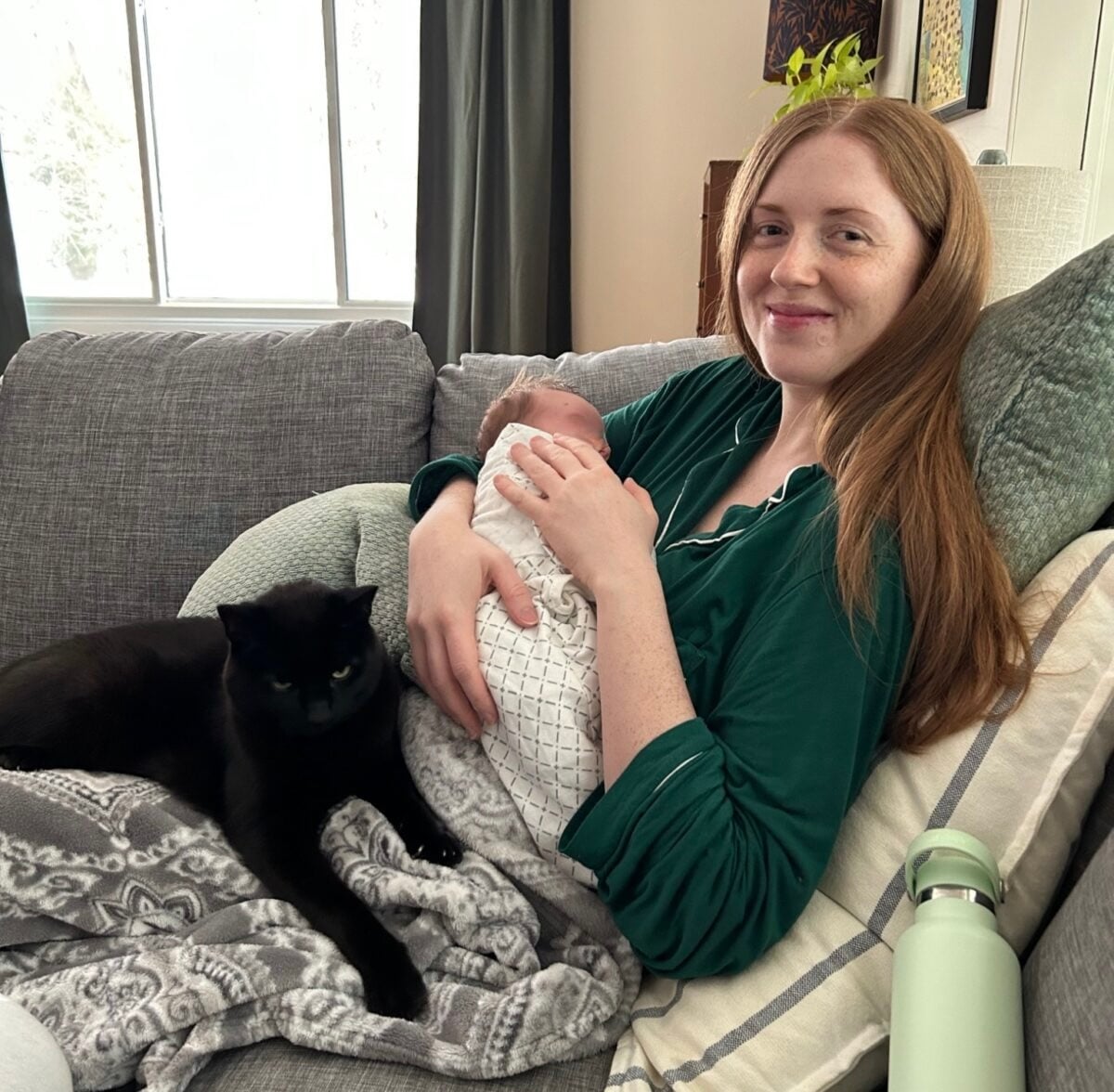 A woman with long red hair sits on a gray couch, holding a swaddled baby. A black cat rests on her lap. There are pillows, a blanket, and a green water bottle nearby. Natural light comes through a window in the background.