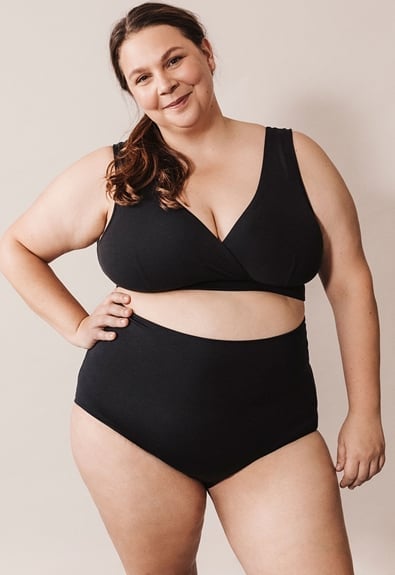 A woman with long brown hair stands against a plain background, wearing a black two-piece swimsuit. She is smiling, with one hand on her hip and her body slightly angled to the side.