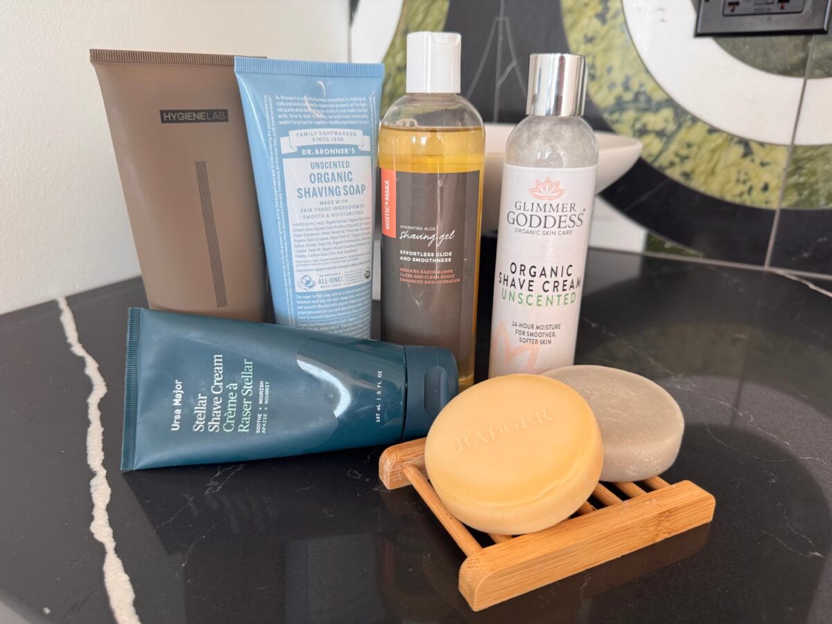 Five shaving and skincare products are arranged on a black countertop, including two bars of soap on a bamboo tray, three bottles/tubes standing upright, and part of a green decorative object visible in the background.