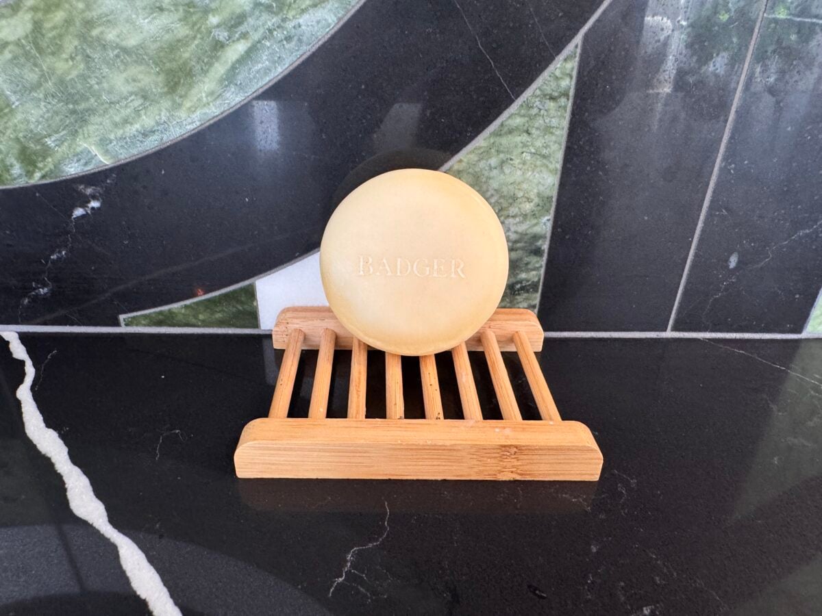 A round bar of soap labeled BADGER sits on a wooden soap dish with slats. The soap and dish are placed on a black, green, and white tiled surface.