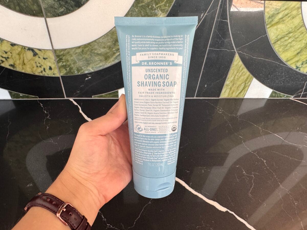 A hand holds a tube of Dr. Bronners Unscented Organic Shaving Soap against a black and green marble-patterned background. The product label is clearly visible.