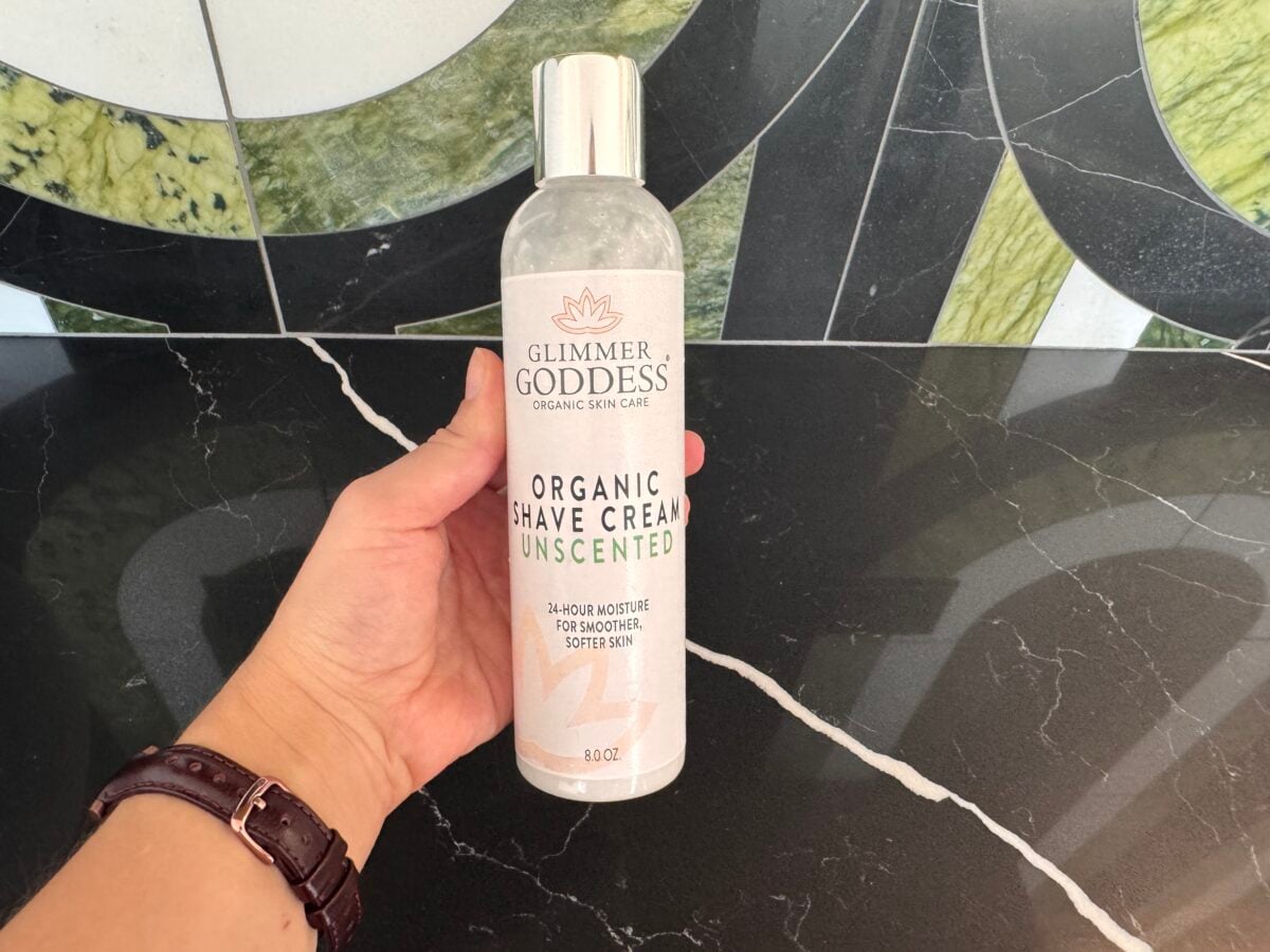 A hand holds a bottle of Glimmer Goddess Organic Shave Cream Unscented in front of a black and green marble-patterned wall. The bottle label mentions 24-hour moisture and softer skin.