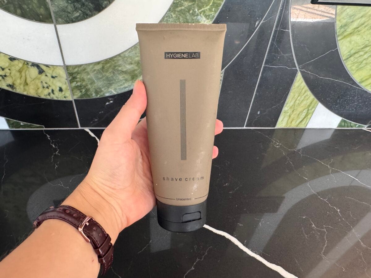 A hand holds a tan tube labeled HYGIENELAB shave cream unscented over a black marble countertop with a geometric green, white, and black tiled backsplash in the background.