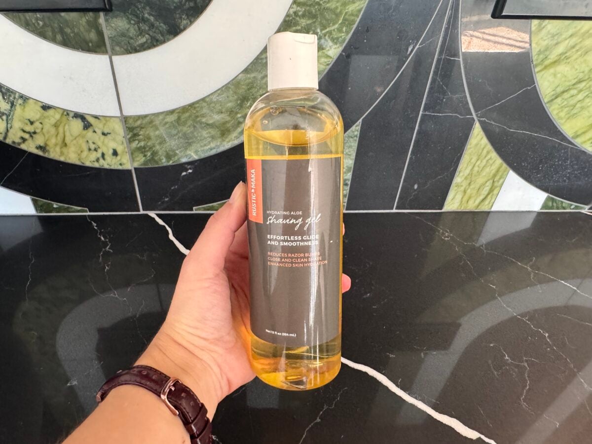 A hand holds a bottle of orangish translucent liquid labeled Effortless Cleanse and Smooth against a black marble countertop and a mosaic tile backsplash with green, white, and gray patterns.