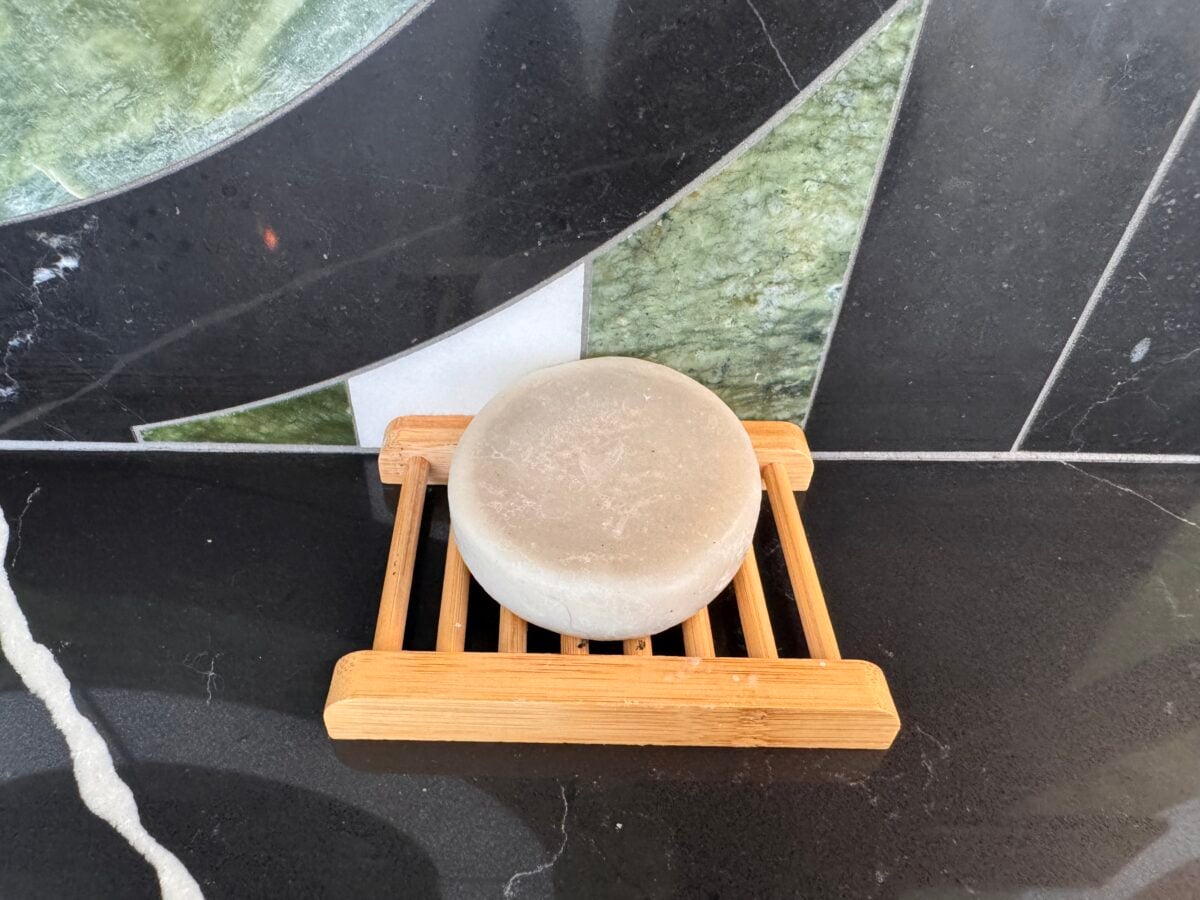 A round, beige bar of soap sits on a small, rectangular wooden soap dish with slats, placed on a black countertop in front of a tiled wall with green, black, and white geometric patterns.