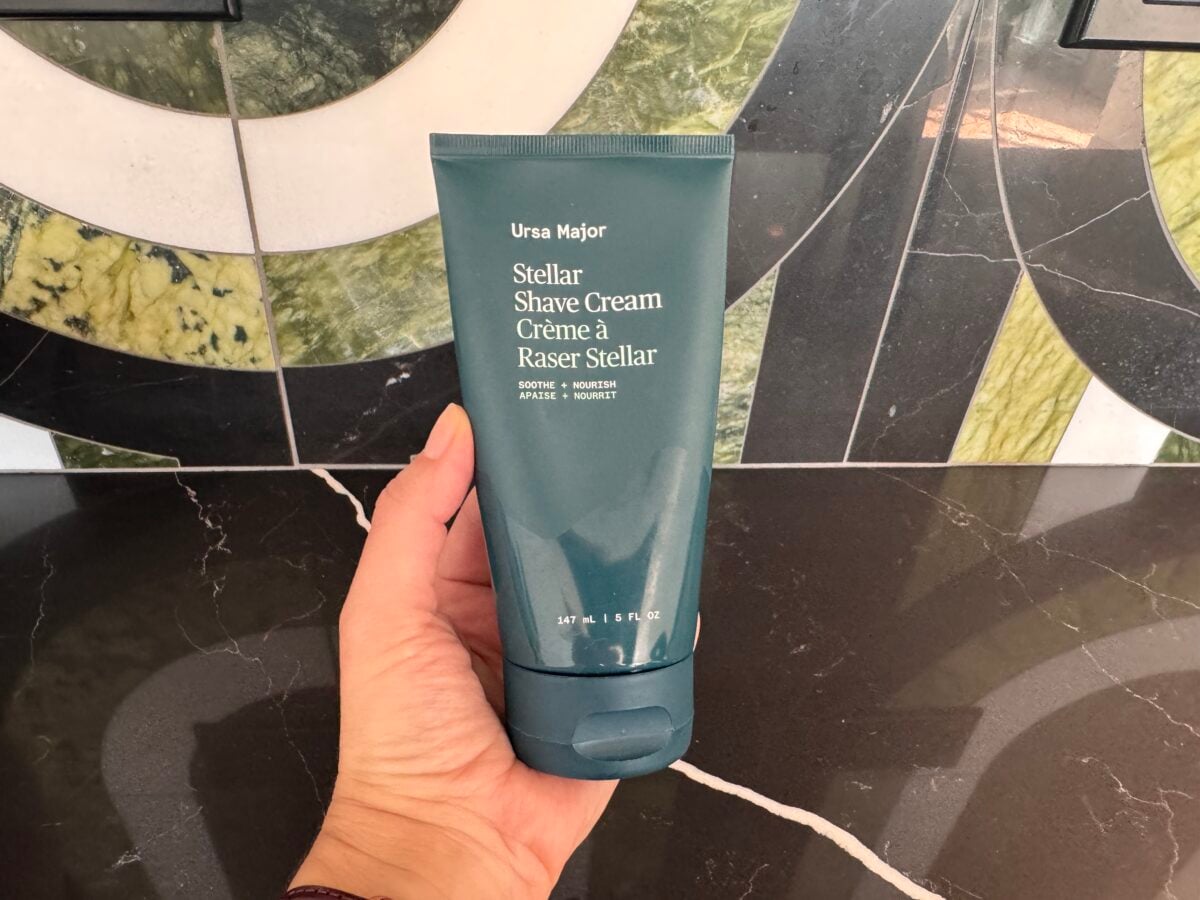 A hand holds a blue-green tube of Ursa Major Stellar Shave Cream against a patterned tile background with green, white, and black colors. The tube is upright and the product label is clearly visible.