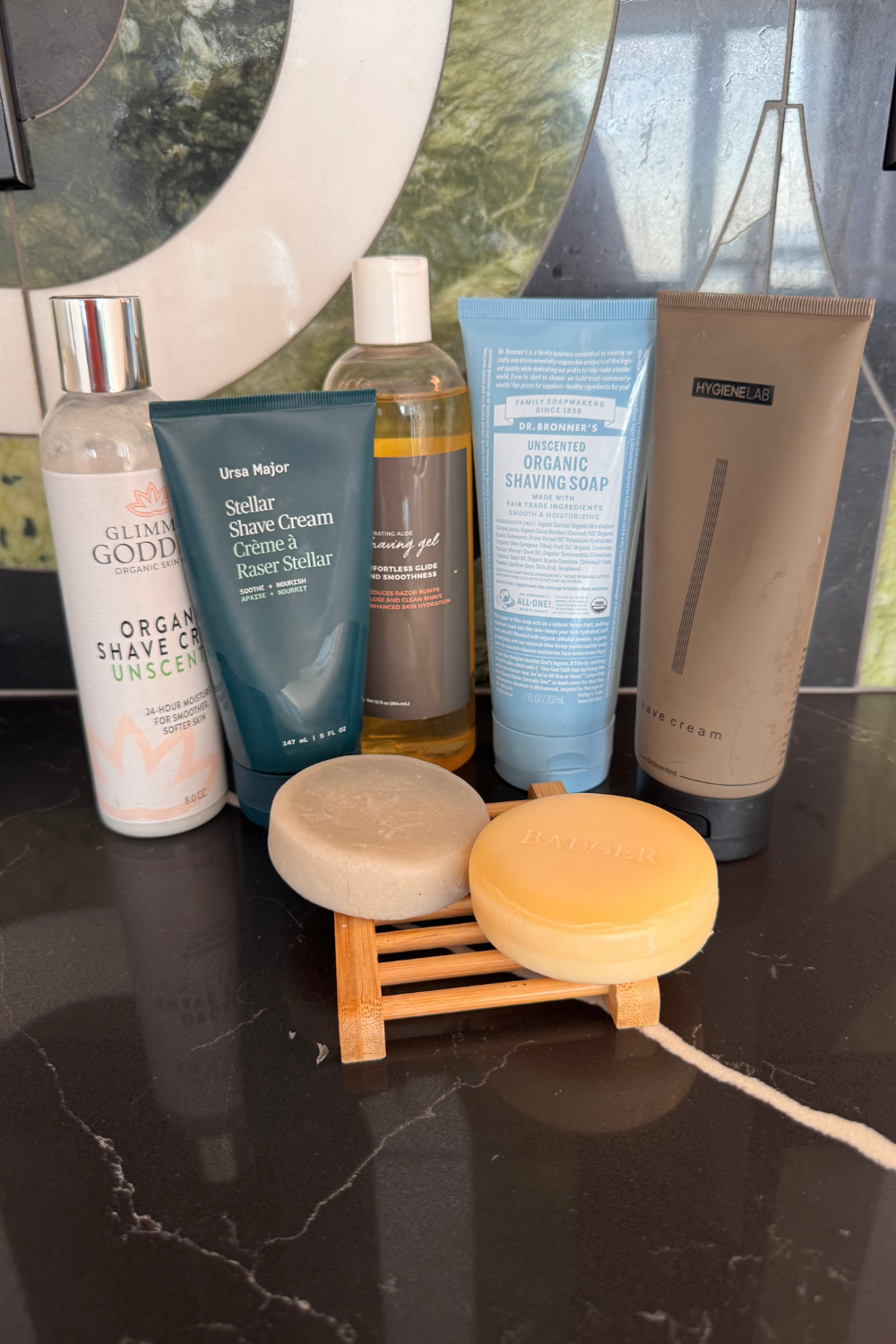 Five bottles and tubes of shaving cream and gel, along with two round soap bars on a wooden soap dish, are arranged on a dark, veined countertop. The products are from brands including Glimmer Goddess, Ursa Major, and Dr. Bronners.