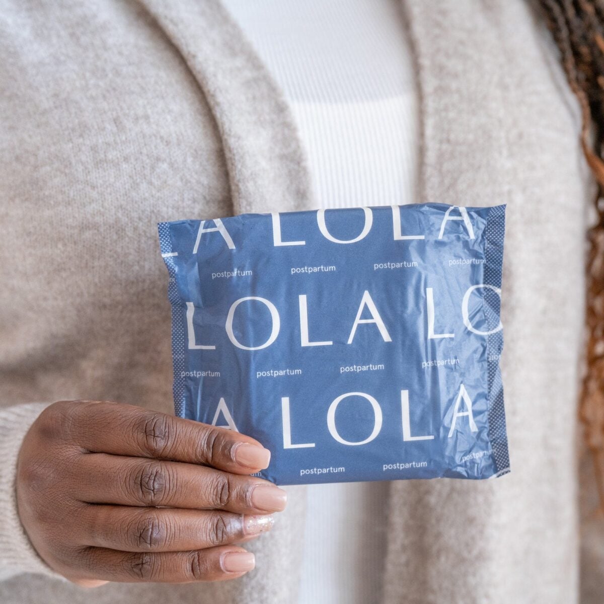 A person wearing a beige sweater holds a blue package labeled LOLA postpartum. The focus is on the hand and package, with the persons torso visible in the background.