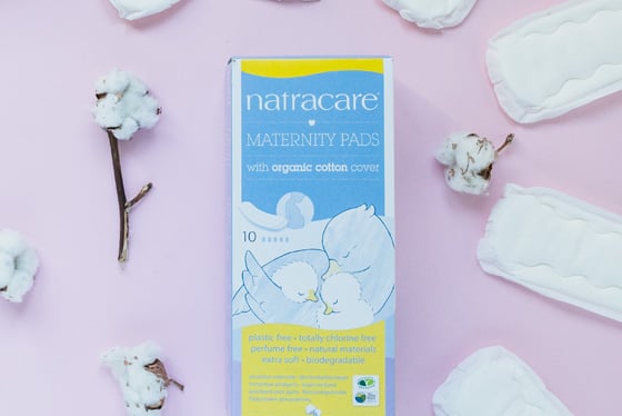 A box of Natracare maternity pads with organic cotton cover is on a pink surface, surrounded by cotton branches and individual maternity pads. The box is blue and yellow with an image of two birds.