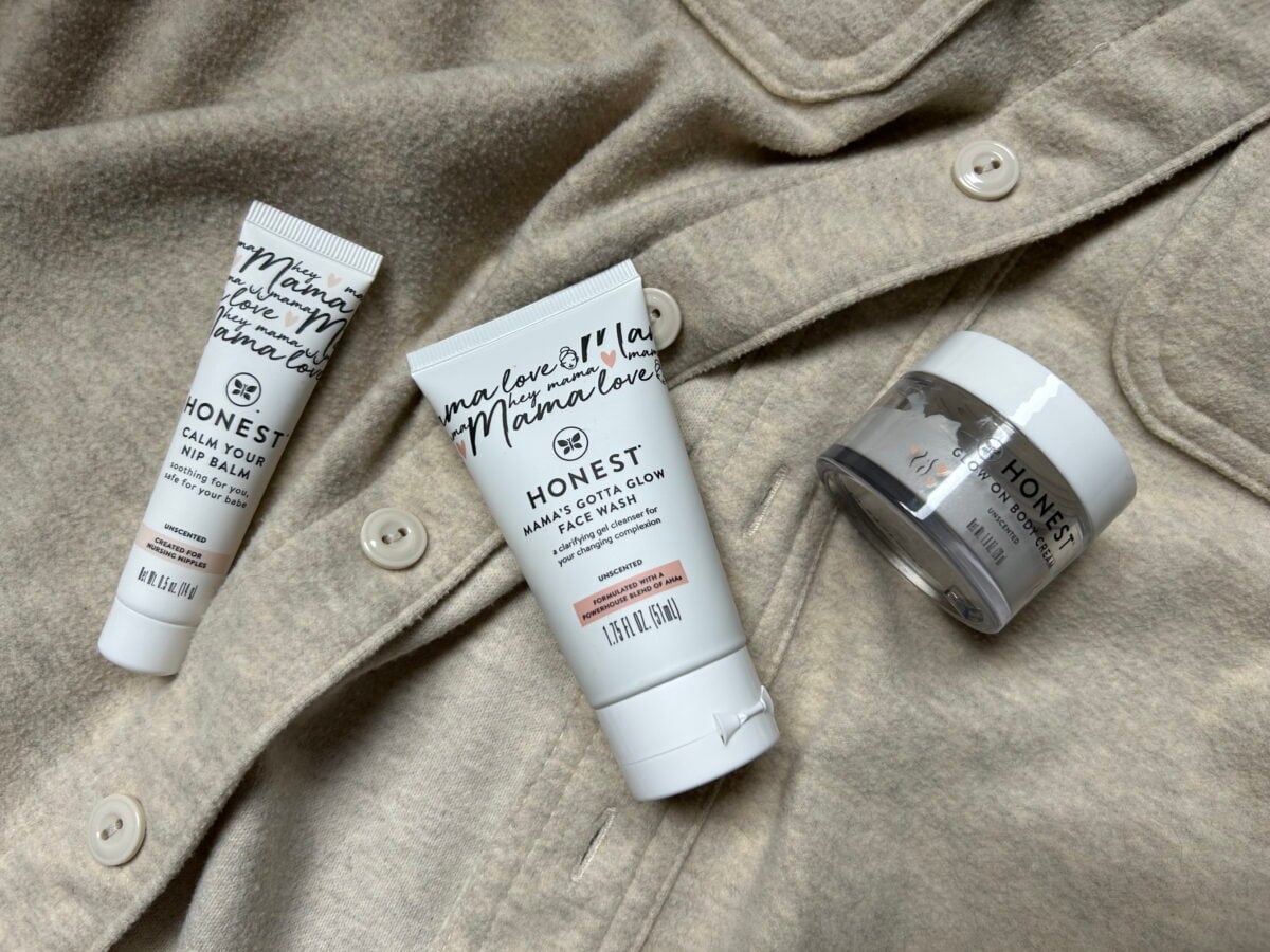 Three Honest beauty skincare products are arranged on a beige button-up garment. The products include a face balm, a face cream, and a small jar, each featuring white packaging with black text.