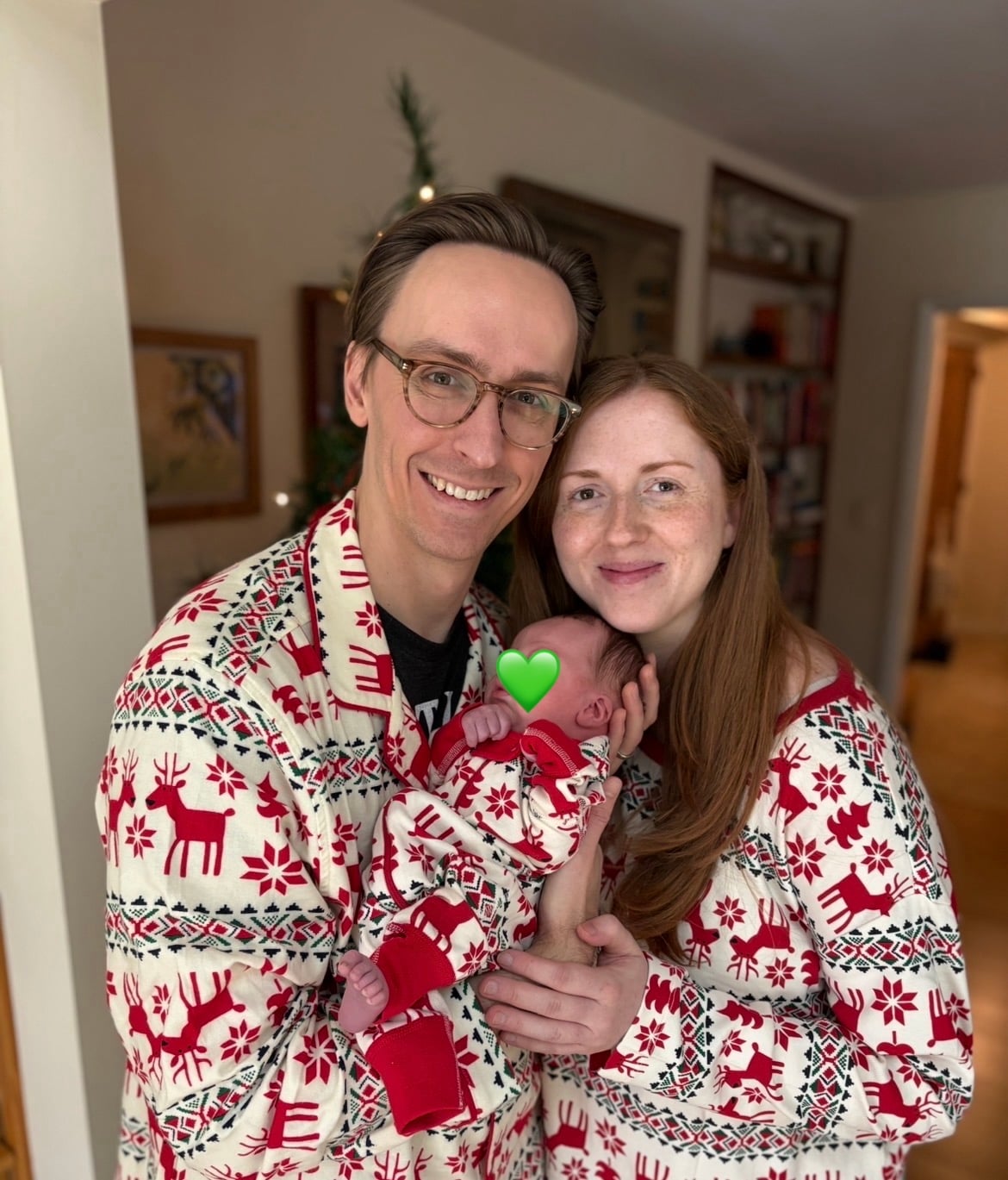 A man and woman smile at the camera while holding a newborn. All three wear matching red and white holiday pajamas with reindeer and snowflake patterns. A green heart emoji covers the babys face.