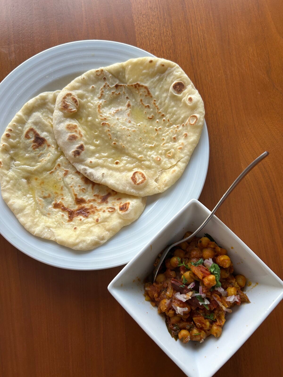 A white plate with two pieces of naan bread next to a square bowl containing chickpea curry garnished with chopped onions and cilantro, placed on a wooden table.