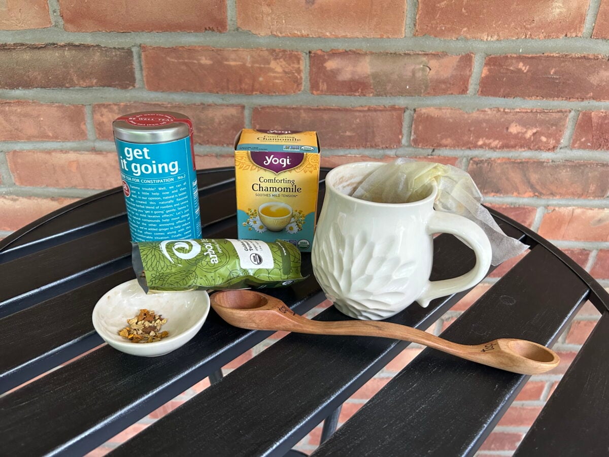 A mug with a tea bag sits on an outdoor table next to a box of chamomile tea, a green tea can, a small dish of loose tea, and a double-sided wooden spoon. Brick wall in the background.
