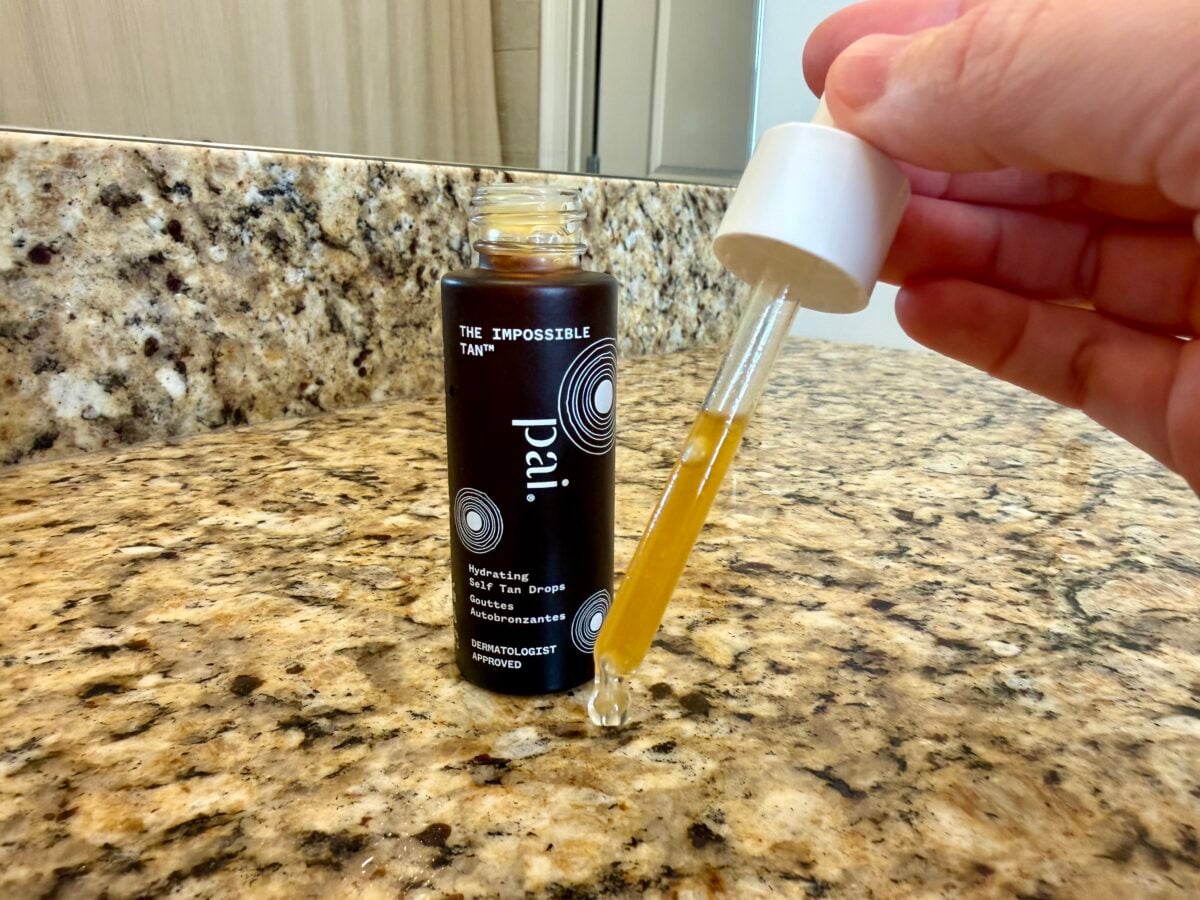 A hand holds a dropper filled with yellow liquid next to an open bottle labeled Pai The Impossible Tan on a granite bathroom countertop.