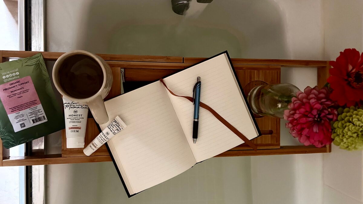 A bathtub tray holds a mug, skincare products, an open notebook with a pen, and a vase with two flowers. The background shows a filled bathtub.