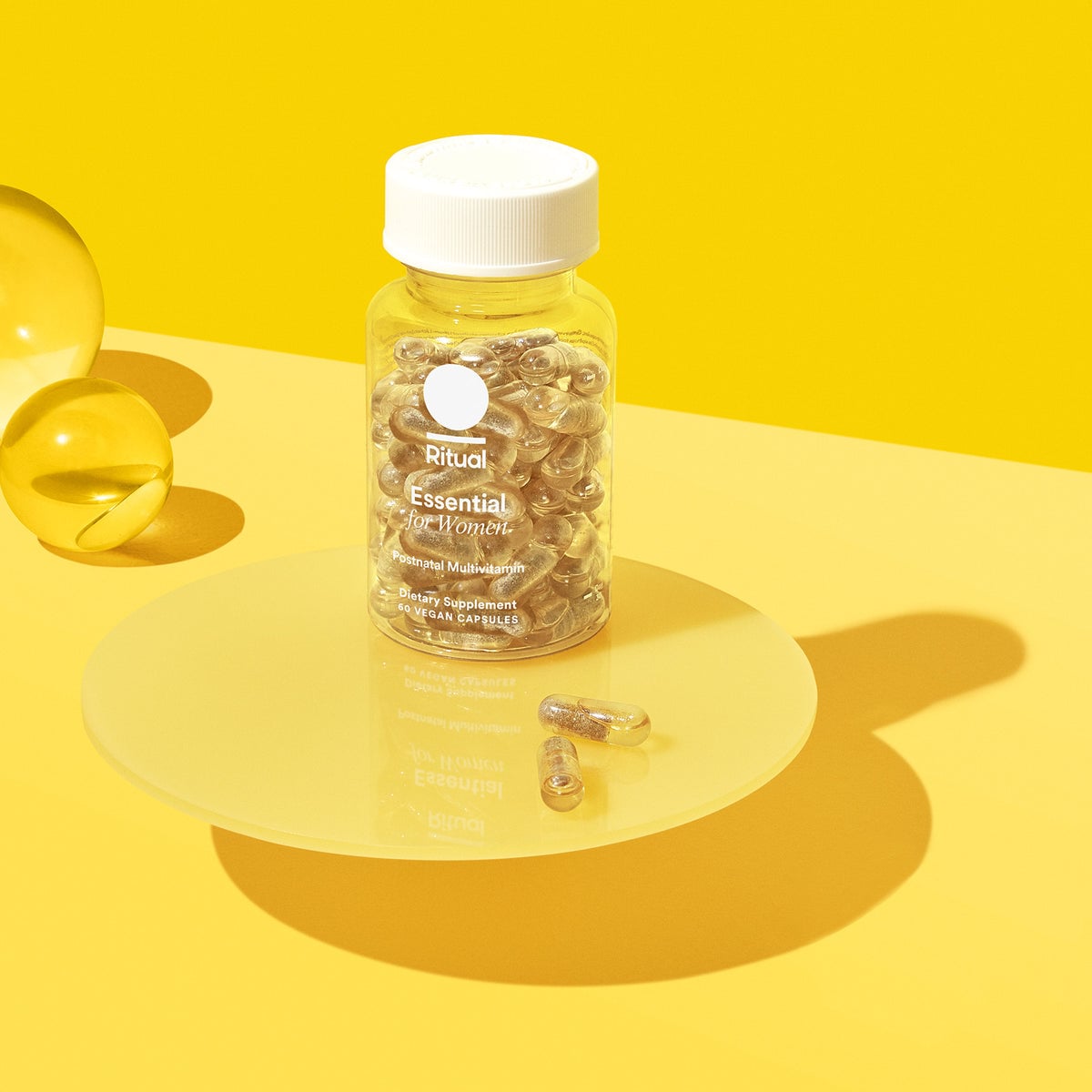 A clear bottle labeled Ritual Essential for Women multivitamin sits on a yellow surface with capsules inside and two loose capsules beside it. The background and lighting are bright yellow.