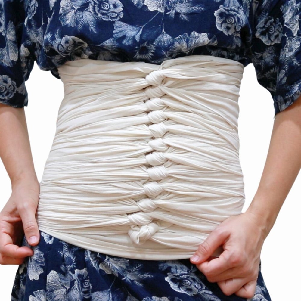 A person wears a blue floral dress and has a long white cloth wrapped and knotted multiple times around their waist, creating a structured, corset-like effect. Only the torso and hands are visible.