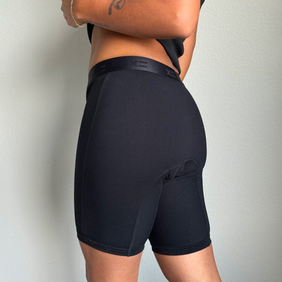 A person is shown from the waist down wearing black fitted shorts and a black top, standing sideways against a plain light-colored wall.