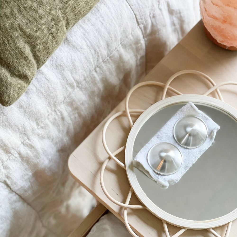 A round mirror on a bedside table holds two metal facial massage tools on a cloth. The table is beside a bed with a white comforter and green pillow. A pink Himalayan salt lamp is also on the table.