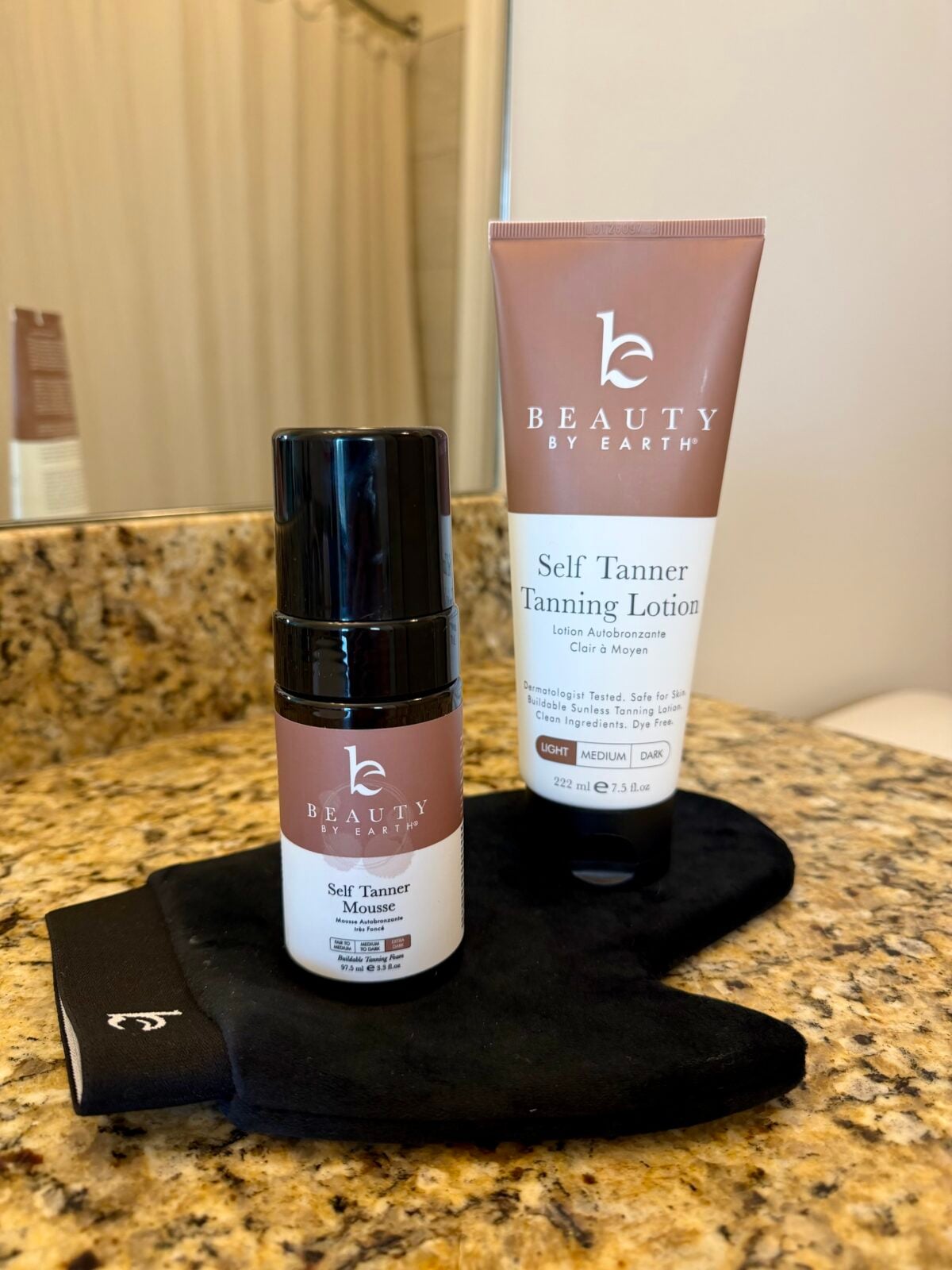 Two Beauty by Earth self-tanning products—a mousse and a lotion—sit on a black tanning mitt on a granite bathroom countertop, with a mirror and shower curtain visible in the background.