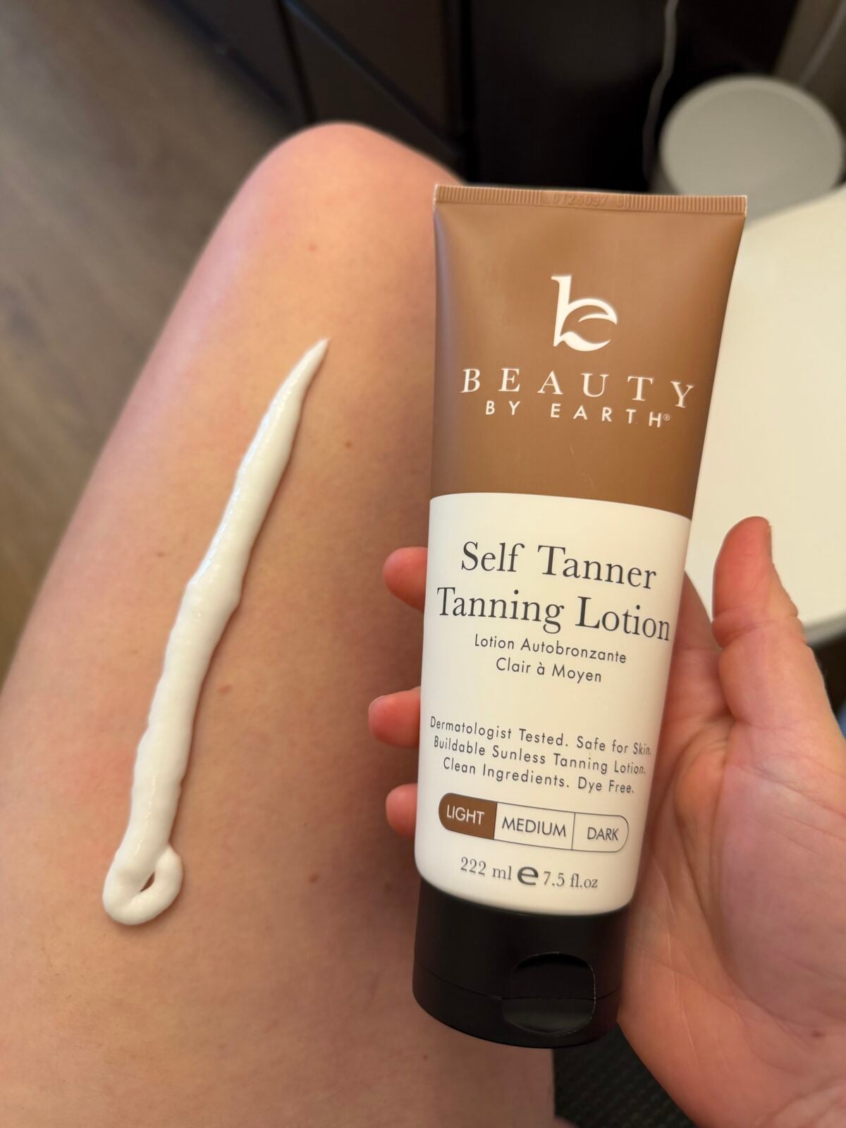 A hand holds a tube of Beauty by Earth Self Tanner Tanning Lotion over a bare leg. A line of white lotion is applied on the leg. The lotion tube label displays product details.
