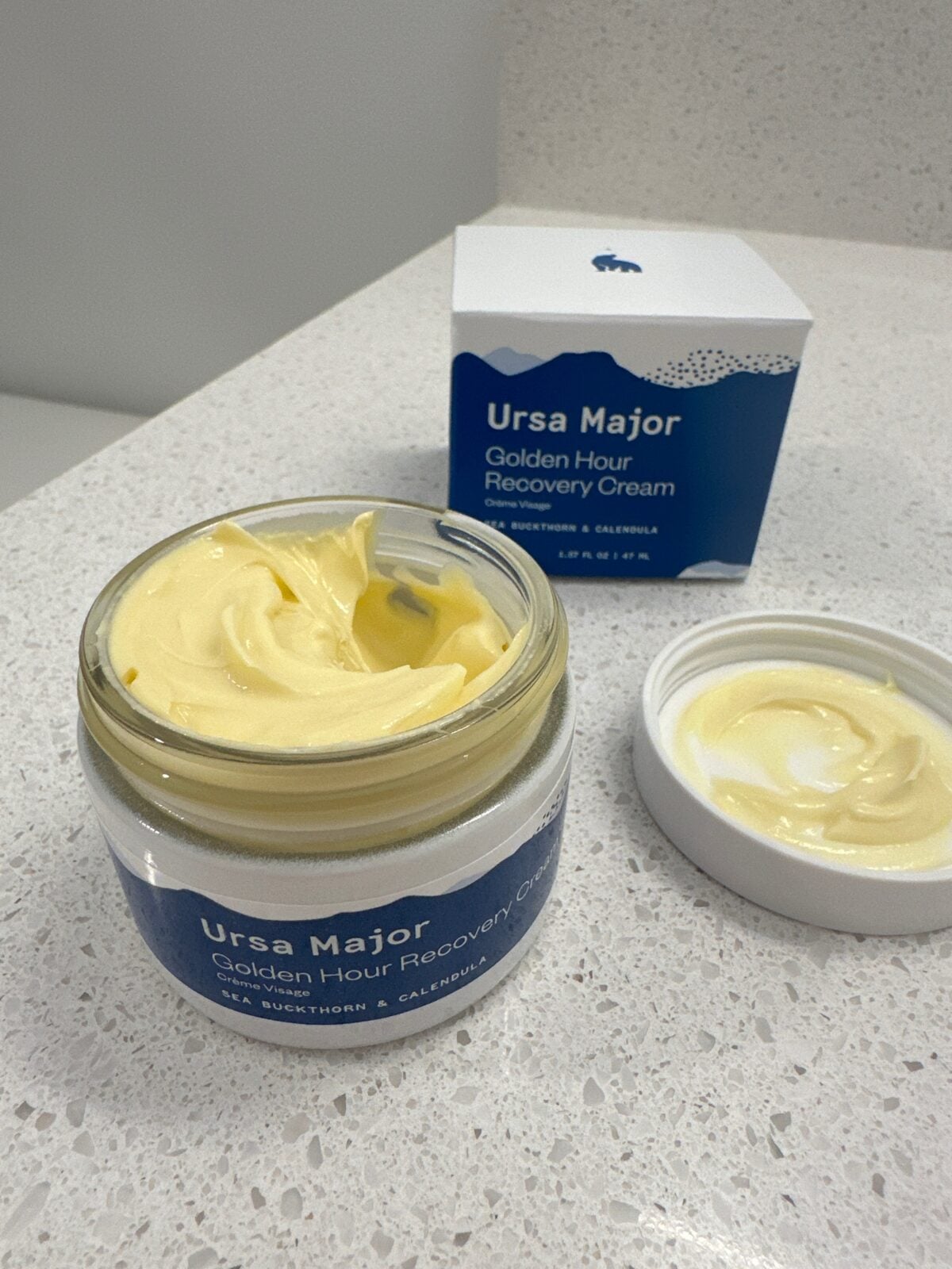 An open jar of Ursa Major Golden Hour Recovery Cream sits on a countertop, with some cream visible on the container lid. The product box is in the background, showing the same label and blue-and-white design.