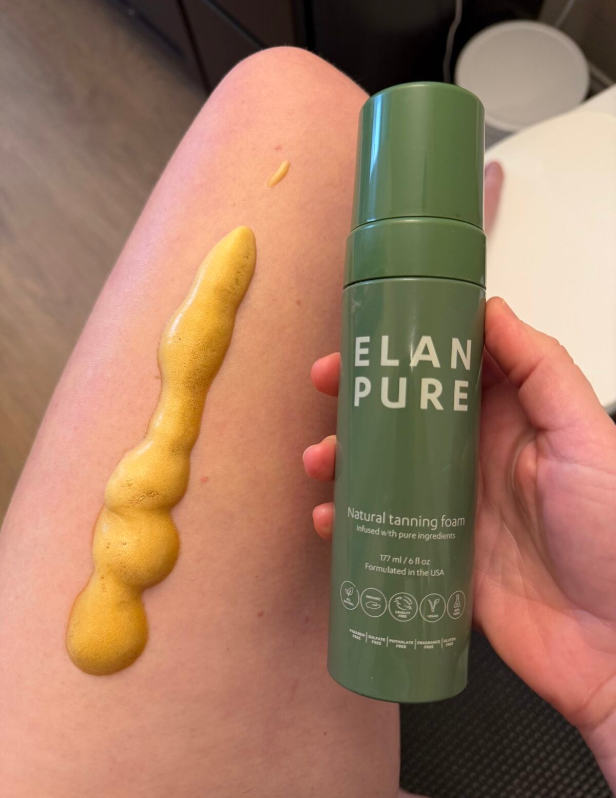 A hand holds a green bottle labeled ELAN PURE Natural tanning foam next to a persons bare thigh, which has a line of yellow-brown tanning foam applied on the skin.