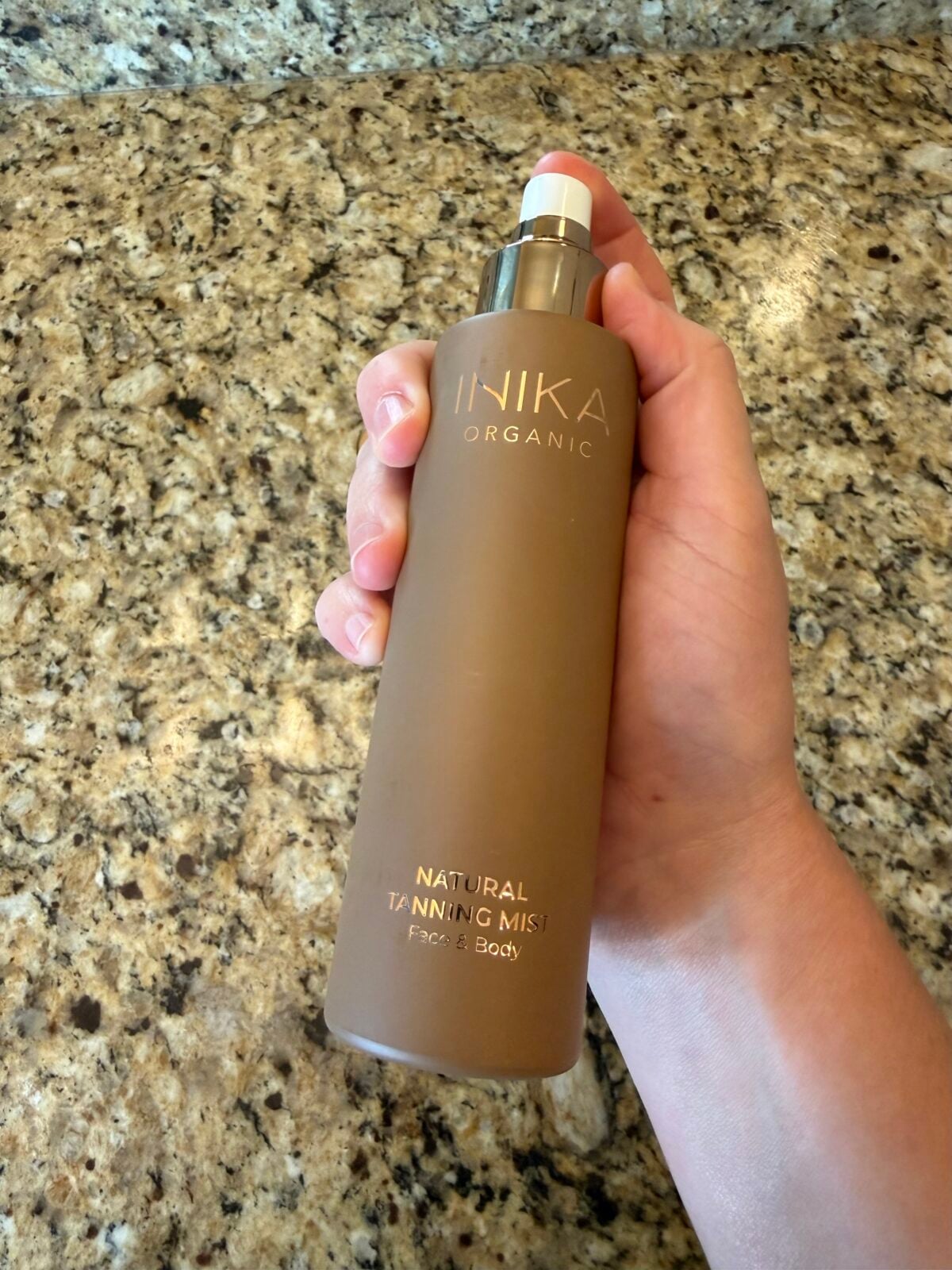 A hand holding a brown bottle labeled Inika Organic Natural Tanning Mist Face & Body against a speckled granite countertop background.