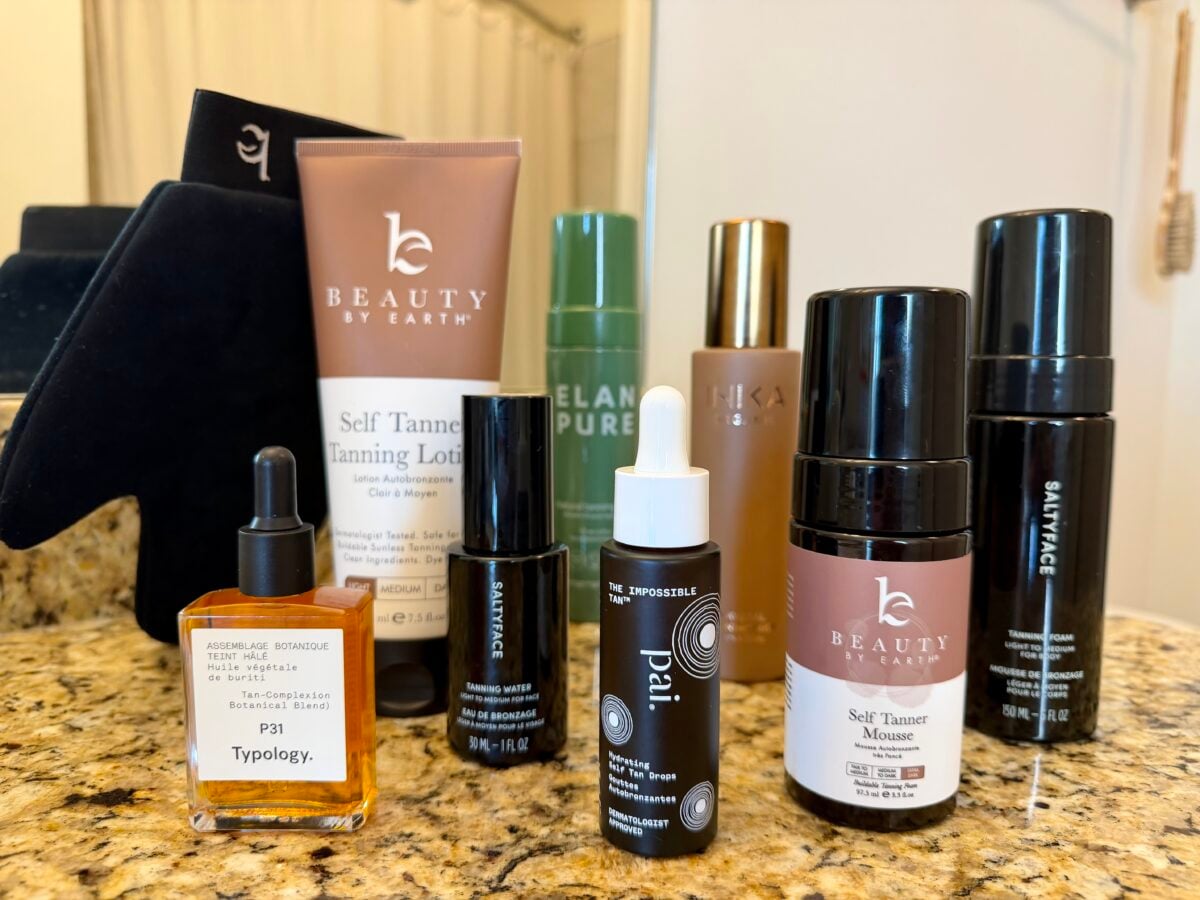 A variety of skincare and tanning products, including lotions, serums, and mousse, are arranged upright on a marble countertop in a well-lit bathroom setting.