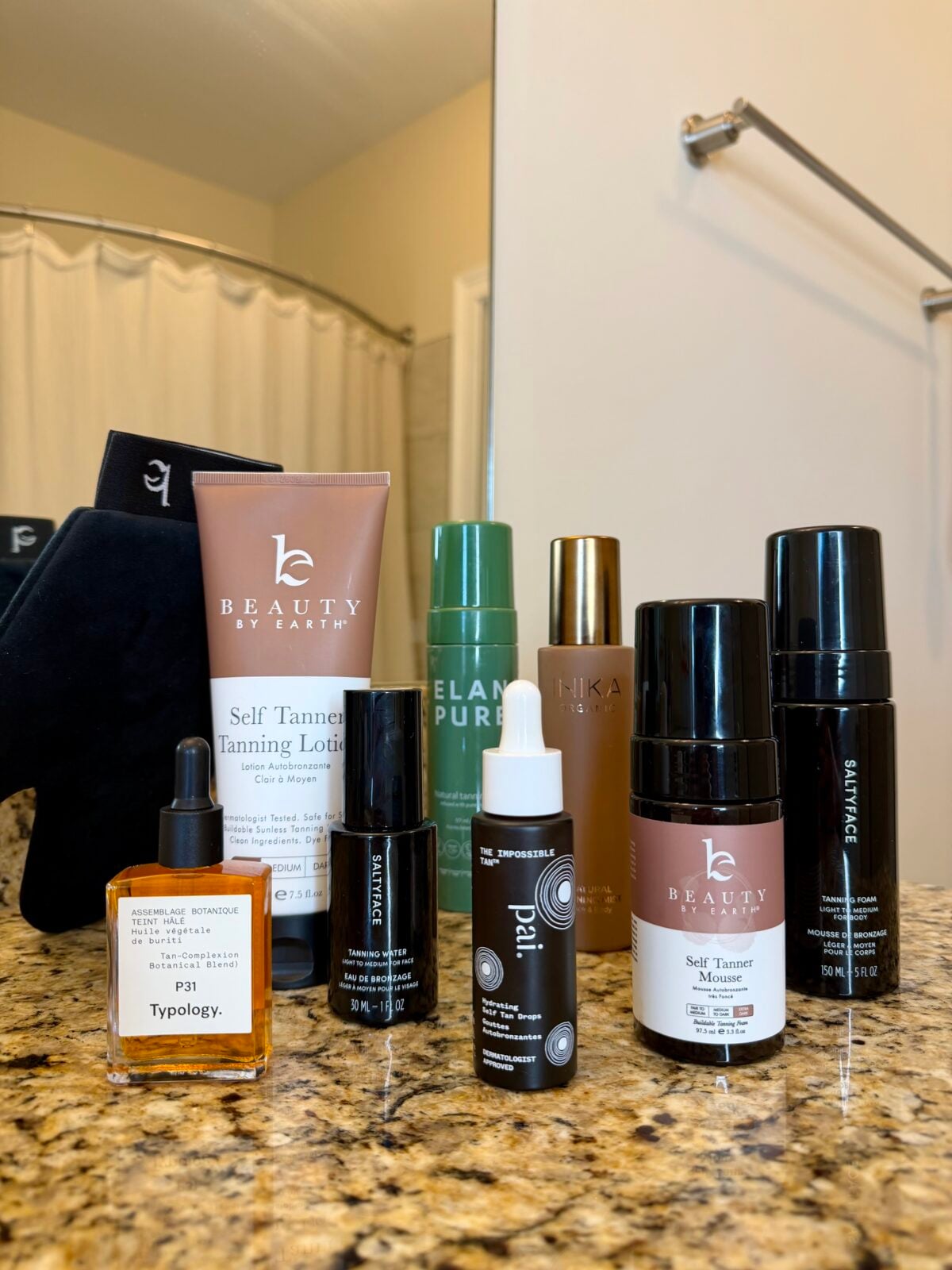 A collection of self-tanning products and serums is displayed on a bathroom counter with a shower and white towels in the background. The items include bottles, tubes, and a dropper bottle.