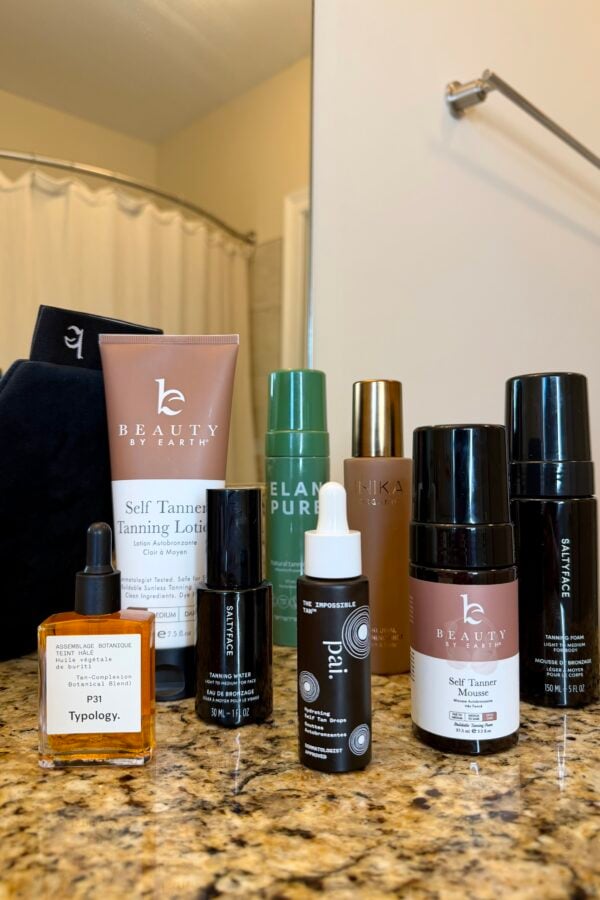 A collection of self-tanning products and serums is displayed on a bathroom counter with a shower and white towels in the background. The items include bottles, tubes, and a dropper bottle.