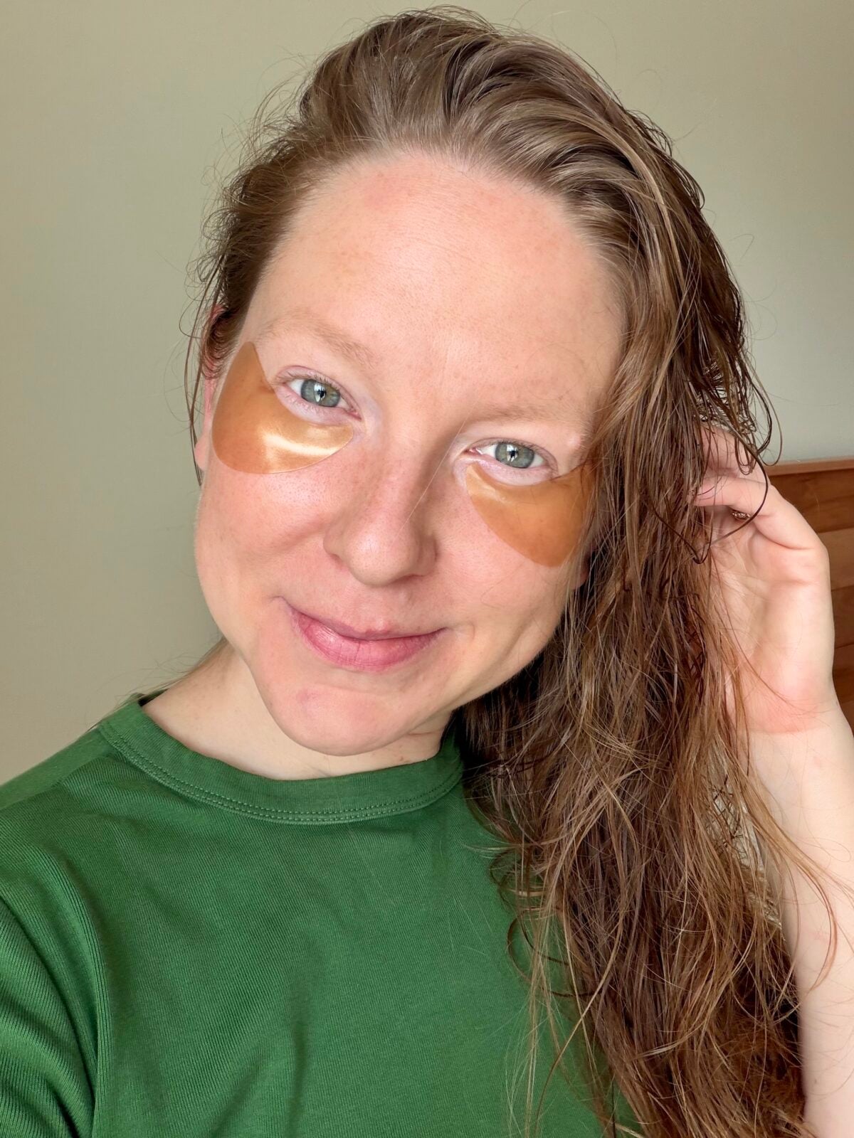 A person with long, wet hair wears a green shirt and gold under-eye patches, smiling at the camera in a well-lit indoor setting.