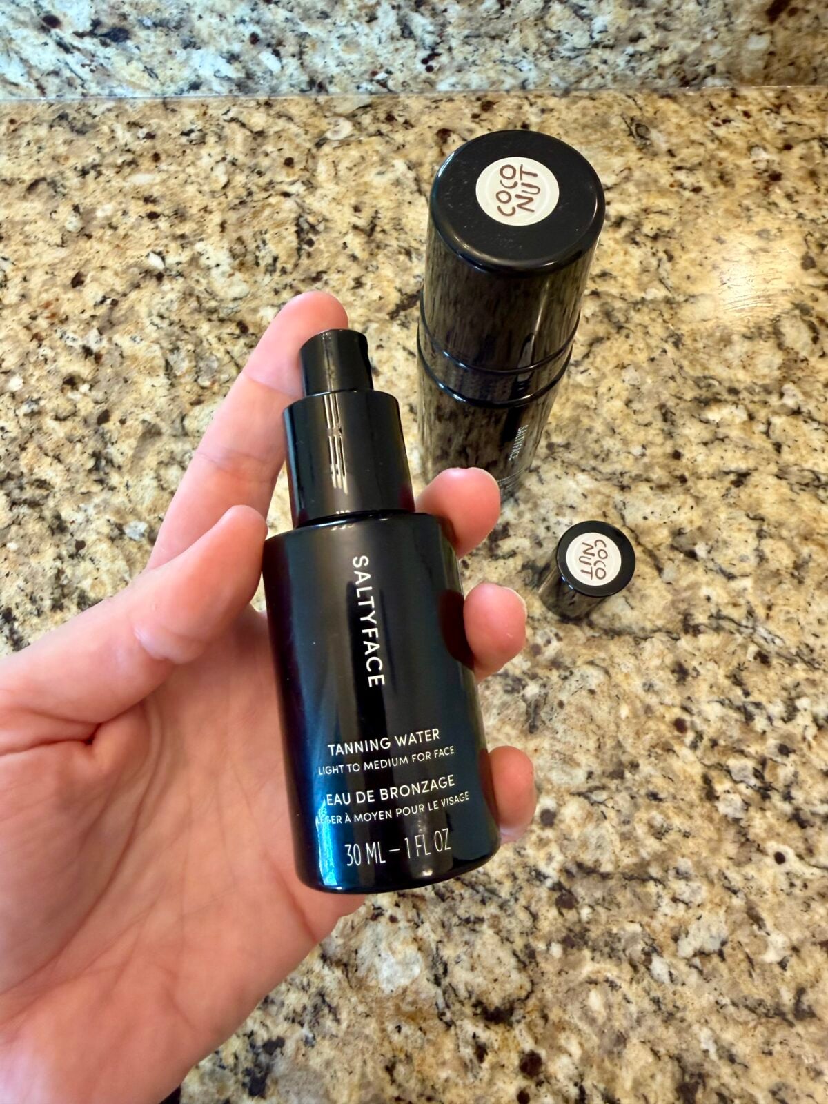 A hand holds a black bottle labeled Saltface Tanning Water above a granite countertop. Nearby, a closed black cylinder and its detached lid are also visible on the counter.