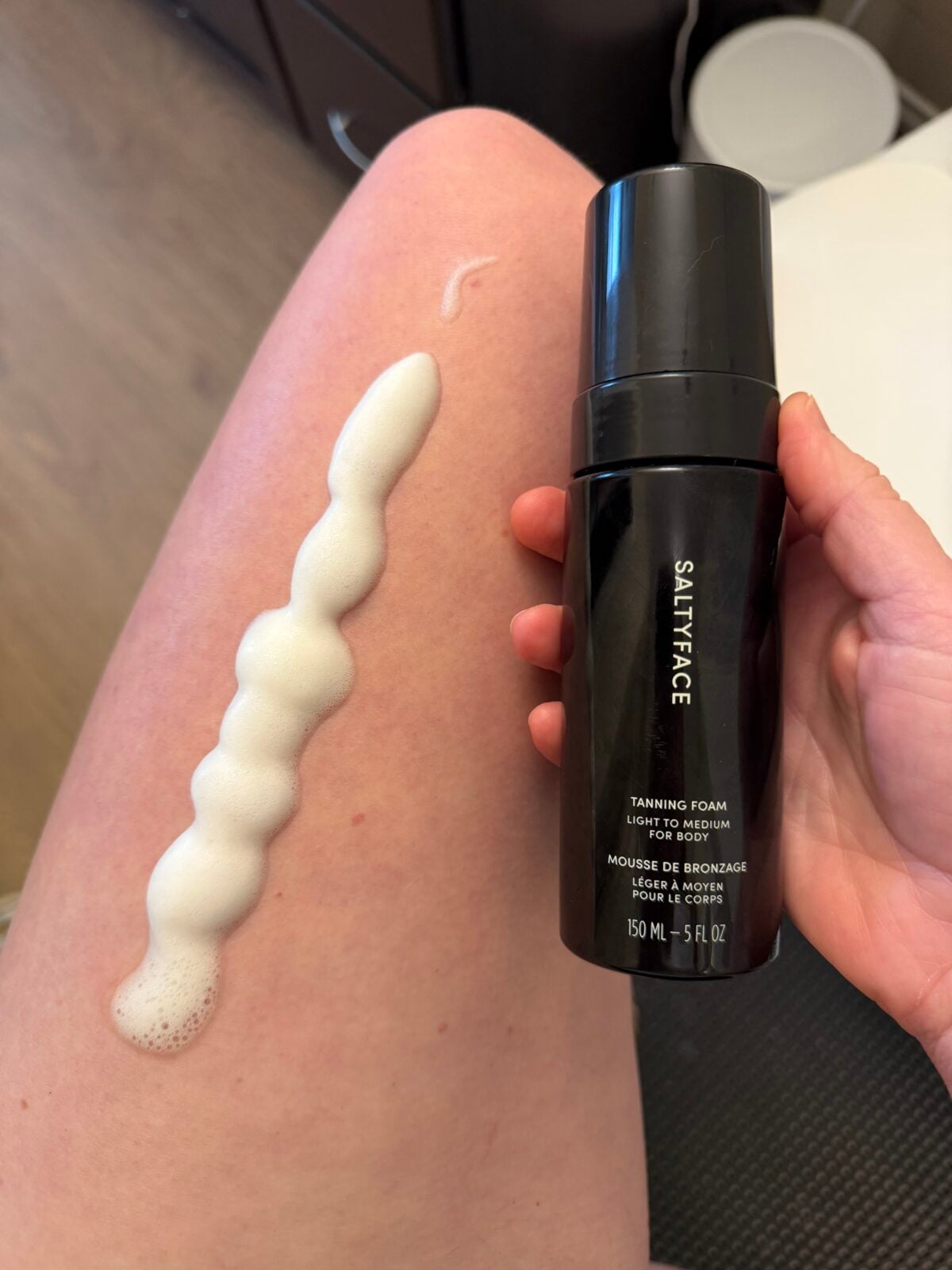 A hand holds a black bottle labeled SALTYFACE Tanning Foam next to a leg with a line of white foam applied to the skin. The bottle contains 150 ml (5 fl oz) of product.