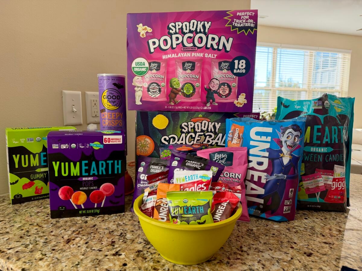 An assortment of organic and spooky-themed Halloween candies and snacks, including boxes of YumEarth pops, a bag of Unreal chocolate bars, and a variety pack of LesserEvil popcorn, displayed on a kitchen counter.