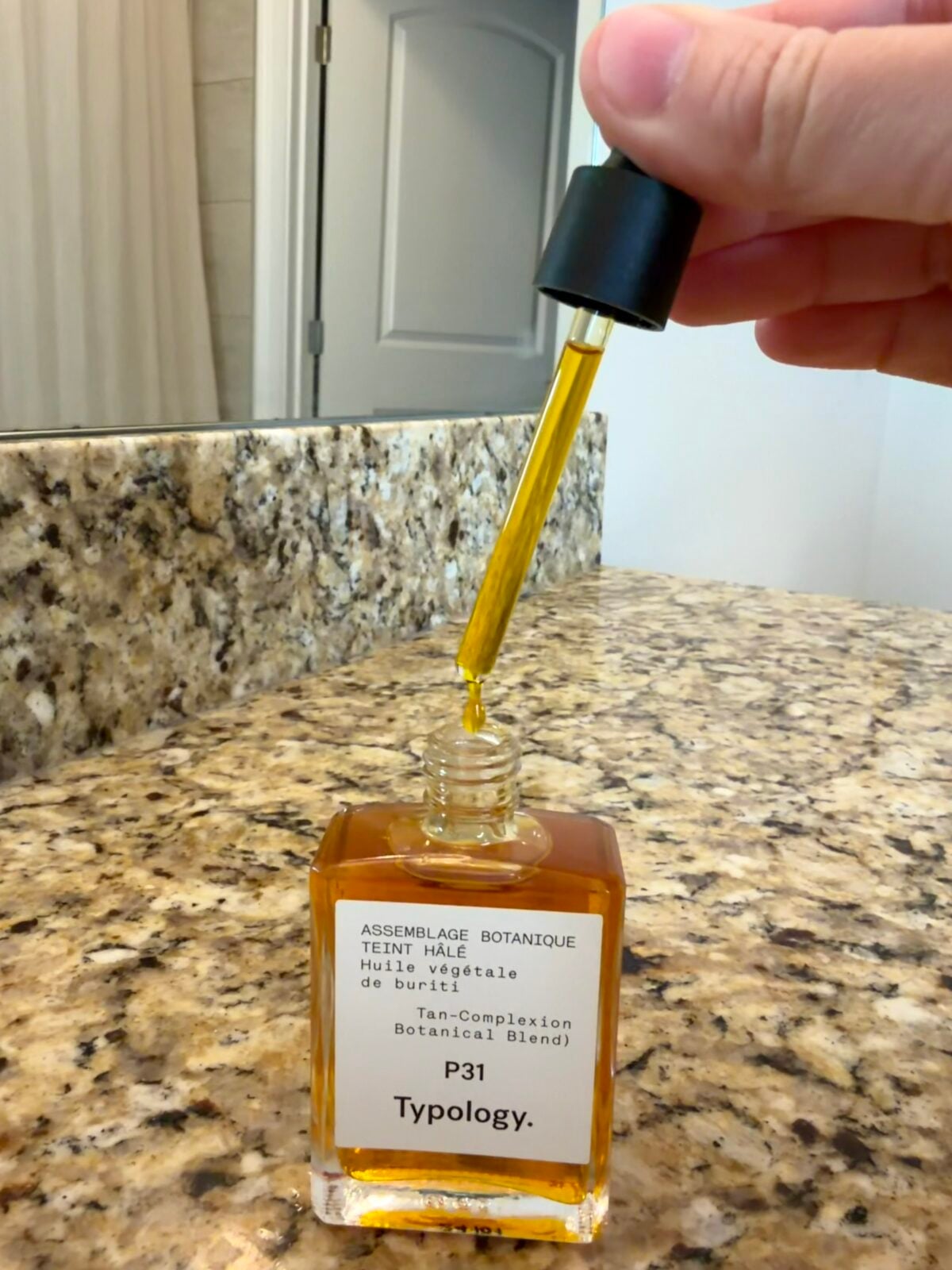 A hand holds a dropper above an open glass bottle labeled Typology P31 Tan-Complexion Botanical Blend, filled with amber-colored oil, on a granite bathroom countertop.