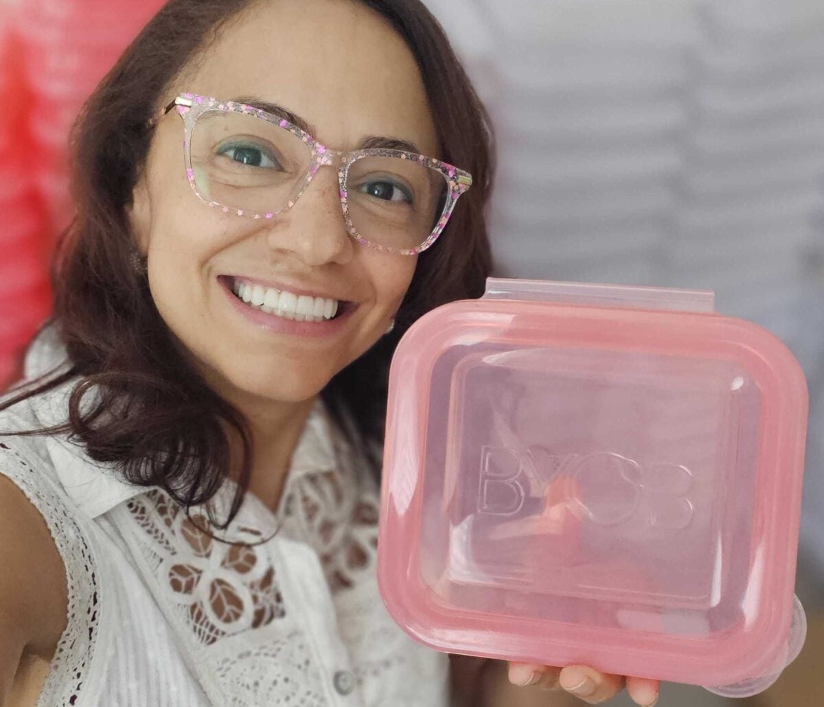 A woman wearing patterned glasses smiles while holding a pink plastic food container embossed with the letters BYOB.