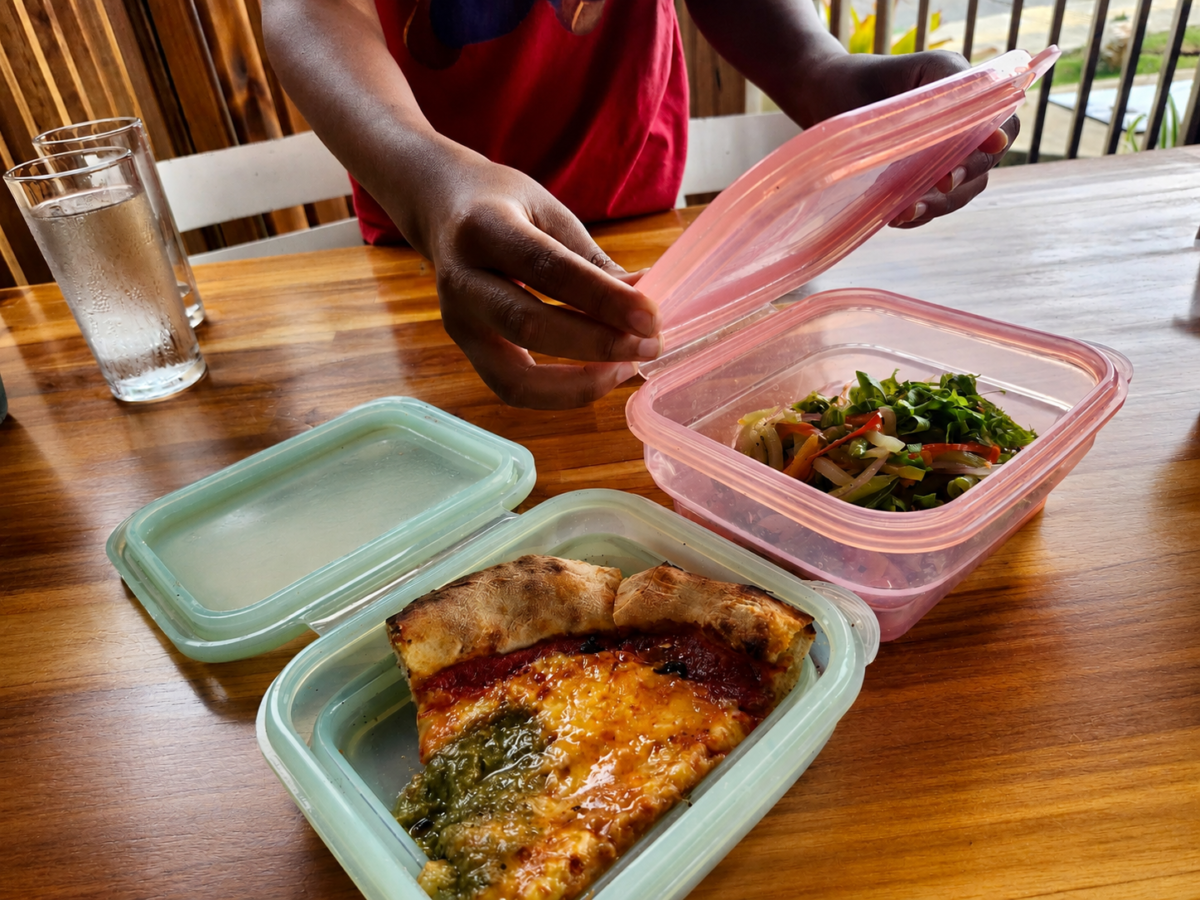 A person opens a pink plastic container of salad next to a light green container holding a slice of pizza on a wooden table.