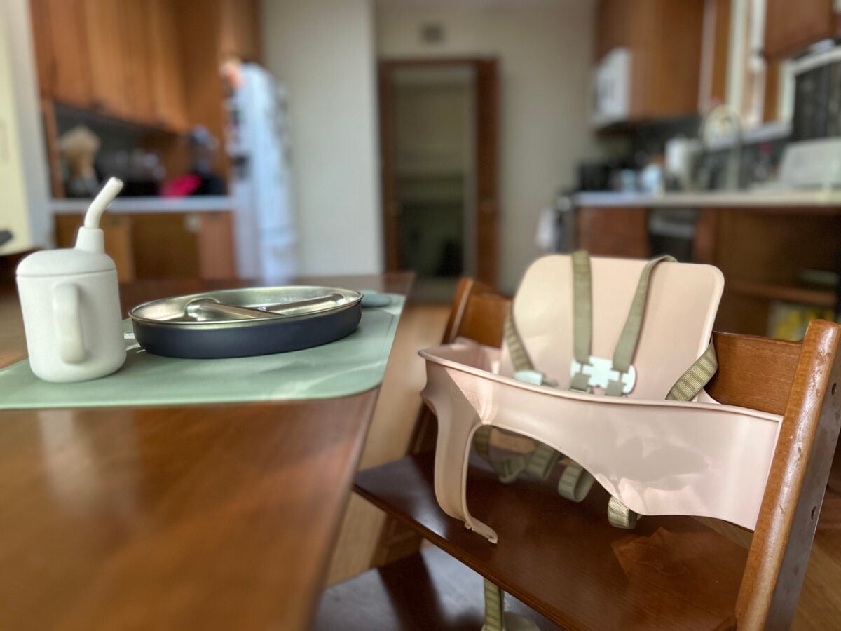 A pink high chair pushed up to a wooden dining table set with a toddlers white cup and a metal tray.