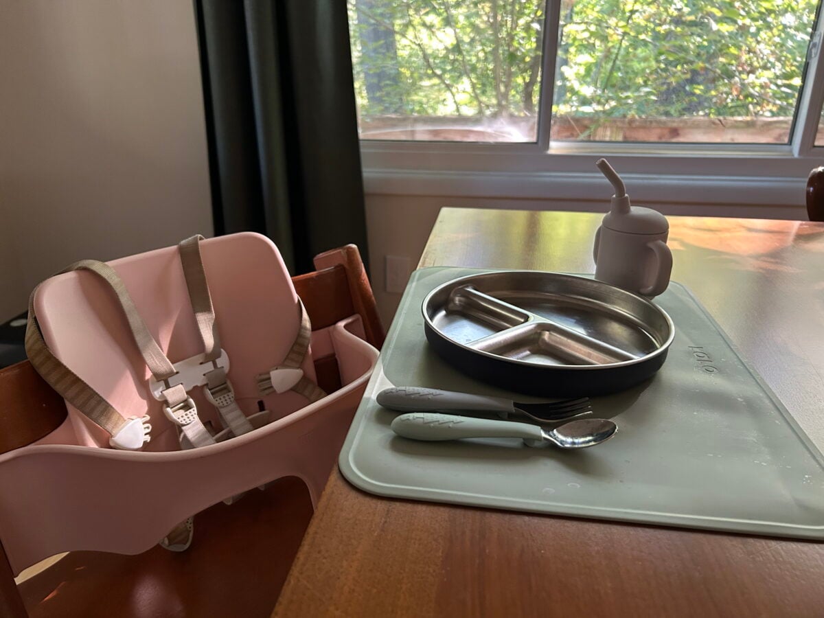 A pink high chair next to a wooden dining table set with a divided metal plate, a fork, a spoon, and a small training cup on a silicone mat.