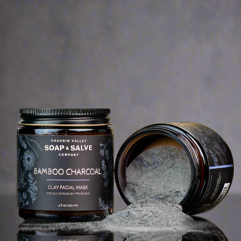 Two jars of Chagrin Valley Soap & Salve Company bamboo charcoal clay facial mask, one upright and one tipped over with gray powder spilled in front.